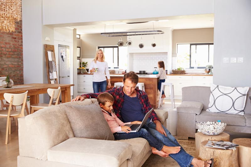 Father and son on couch reading a book while rest of family is in kitchen.